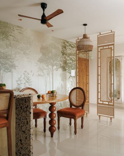 Round wood dining table with cane chairs beneath forest mural, wooden ceiling fan and carved screen partition.