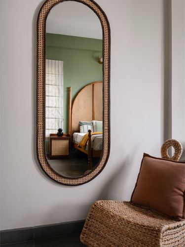 Oval cane-framed mirror reflecting a green-walled bedroom, beside a woven basket chair with terracotta cushion