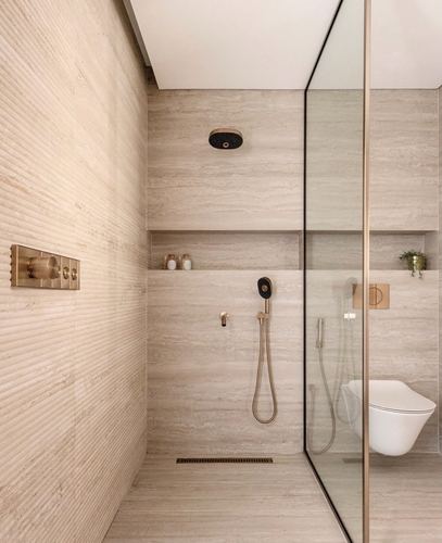 Beige stone-clad walk-in shower with brass fixtures beside a glass-partitioned wall-hung toilet