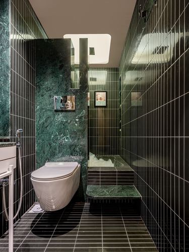 Narrow bathroom with green marble wall, ribbed dark tile, wall-hung toilet, and sunken tub under skylight