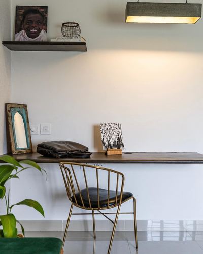 Wall-mounted wood desk with brass wire chair, framed portrait on shelf above, plant in foreground