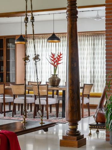 Carved wooden column in foreground with dining table, cane chairs, brass swing, and sheer-curtained windows behind
