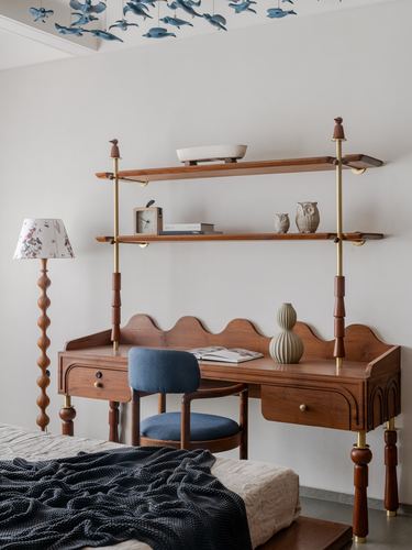 Carved wood desk with brass-poled shelving above, floral-shade lamp, and blue chair in a bedroom
