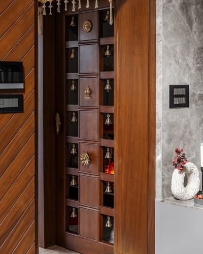 Carved wooden pooja room door with rows of brass bells beside a marble wall and vase