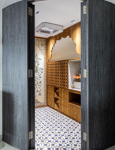 View through curved wooden doors into a pooja room with jali lattice, floral mural, and patterned tile floor