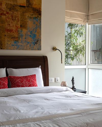 Bedroom corner with carved wood headboard, red embroidered cushions, abstract painting, and brass swing-arm sconce beside a window with greenery.