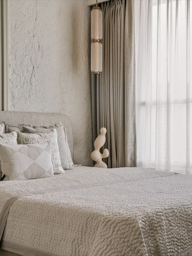 Neutral bedroom corner with textured plaster wall, layered cushions, sheer curtains and a ribbed pendant lamp