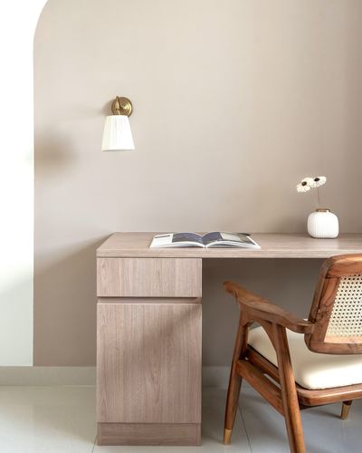 Wooden desk with cane armchair, brass sconce on arched taupe wall, vase of flowers