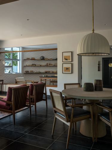Round dining table with cane chairs beneath a large dome pendant, opening to a reading nook with bookshelves.