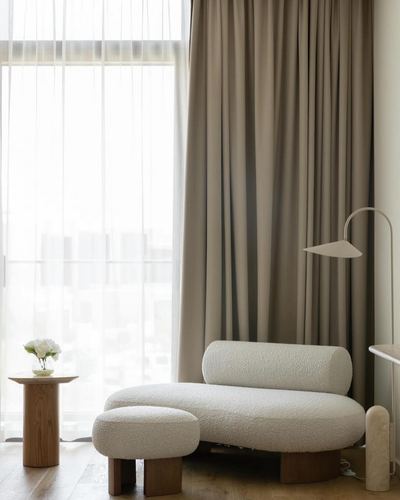 Cream boucle chaise and ottoman by a curtained window with a wooden side table and arc lamp