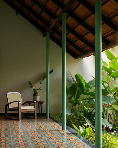 Covered veranda with cane armchair, patterned tile floor, green columns, and lush courtyard plants beyond.