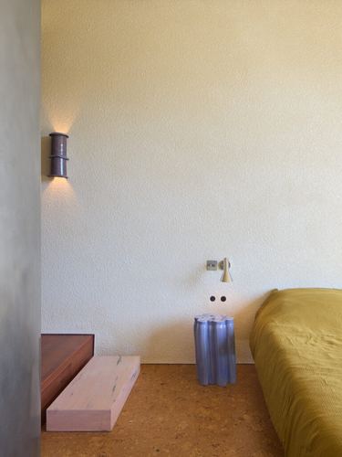 Bedroom corner with textured plaster wall, cylindrical sconce, fluted side table, and mustard bedding on cork floor