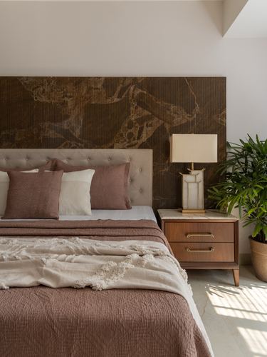 Bedroom with tufted beige headboard, patterned brown wall, wooden nightstand and lamp, plant beside window