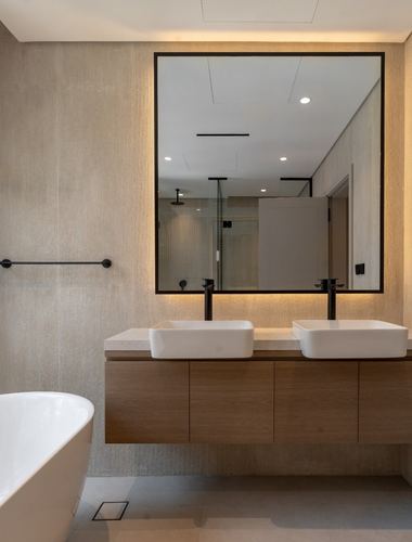 Modern bathroom with dual vessel sinks, wood vanity, backlit square mirror, freestanding tub