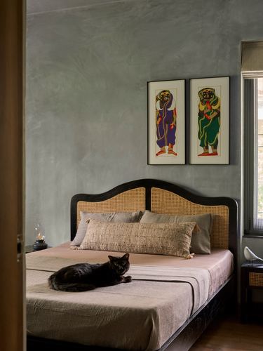 Bedroom with arched cane headboard, textured grey wall, folk art prints, black cat on bed