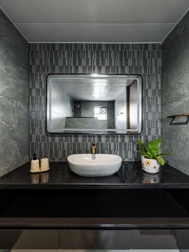 Grey stone bathroom vanity with black countertop, oval white sink, and mirror reflecting corridor beyond