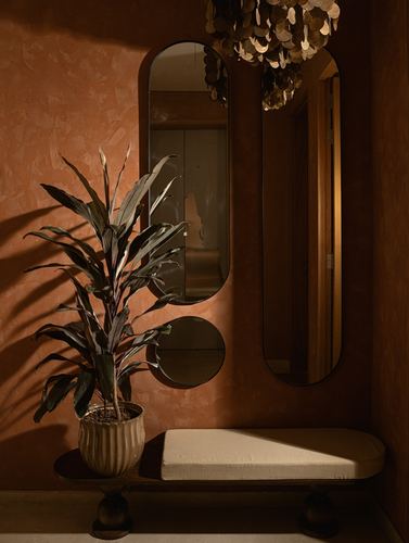Terracotta plastered foyer wall with oval and round mirrors, plant, and upholstered bench