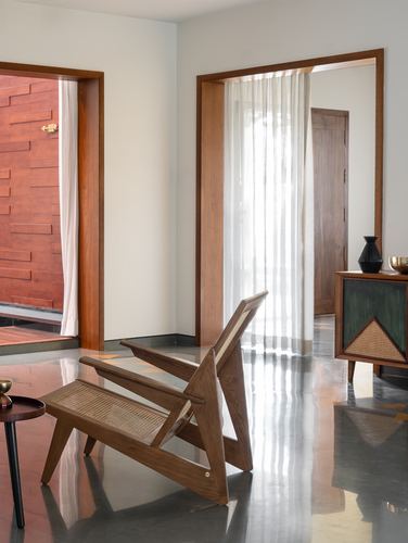Cane and teak lounge chair beside a sheer-curtained doorway, brick wall glimpsed through adjacent opening