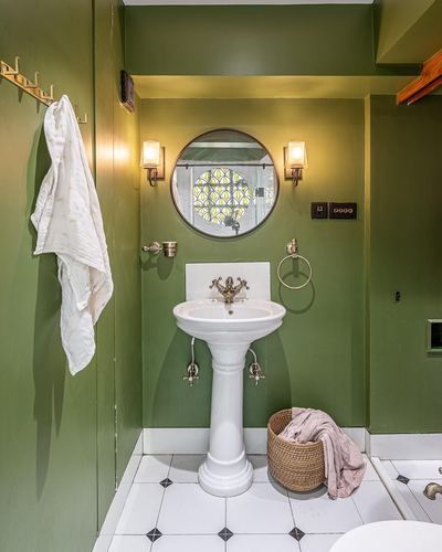 Olive green bathroom with pedestal sink, round mirror, and wicker basket on patterned tile floor