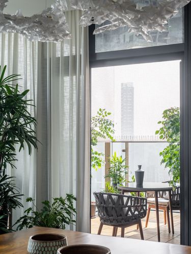 Balcony seen through sheer curtains and glass, framed by potted plants and city skyline