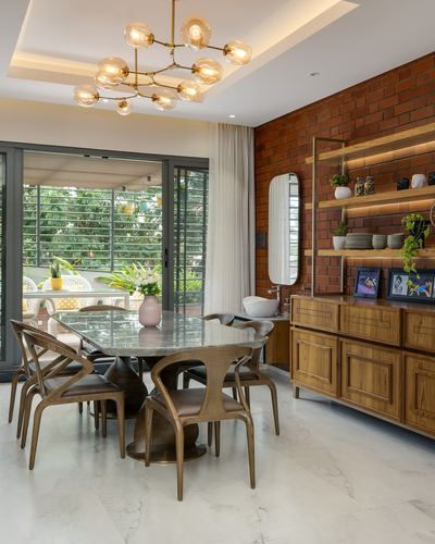Round marble dining table with wood chairs beneath brass chandelier against exposed brick wall