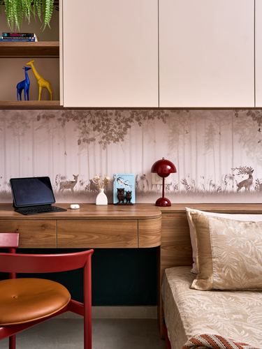 Curved wooden desk with laptop and red lamp against forest-print wallpaper beside a bed