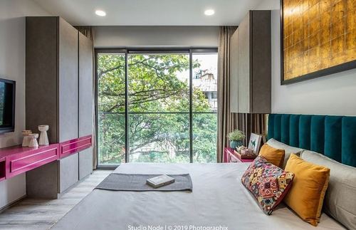 Bedroom with teal velvet headboard, mustard cushions, and floor-to-ceiling window showing trees