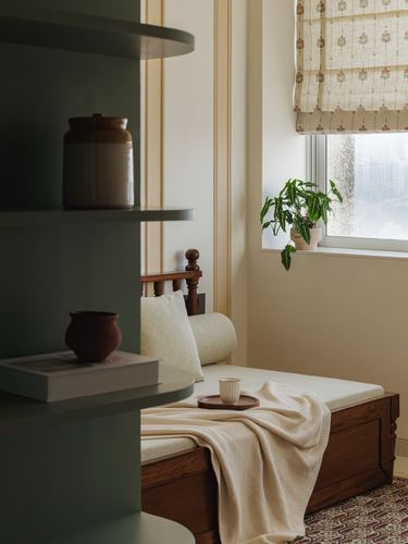 Sage green open shelves beside a wooden daybed with cushions near a window with a patterned blind