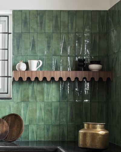 Green glazed tile backsplash with scalloped wood shelf holding ceramics and a brass pot below
