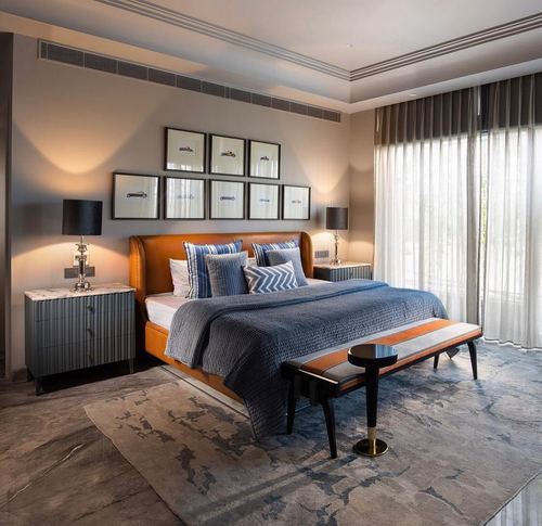 Bedroom with orange leather headboard, blue bedding, gallery wall of car prints, and sheer curtained window.