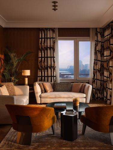 Warm-toned living room with orange velvet chairs, cream sofa, and patterned curtains framing a river view at dusk.