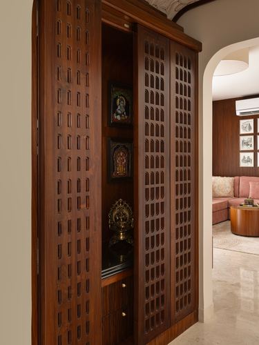 Carved wooden pooja cabinet with lattice doors open, brass idol and framed deities, seen from a passage leading to a living room