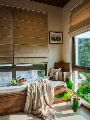 Sunlit window seat corner with woven blinds, cushions, throw blanket and potted plant against greenery view