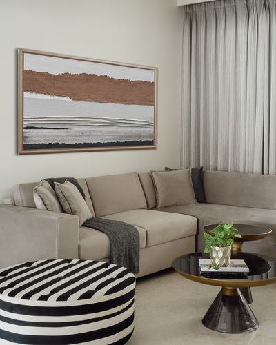 Beige sectional sofa with striped ottoman and round brass coffee table beneath abstract landscape art