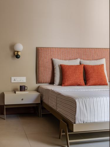 Bedroom corner with striped orange headboard, layered pillows, and a globe wall sconce over a beige bedside table.