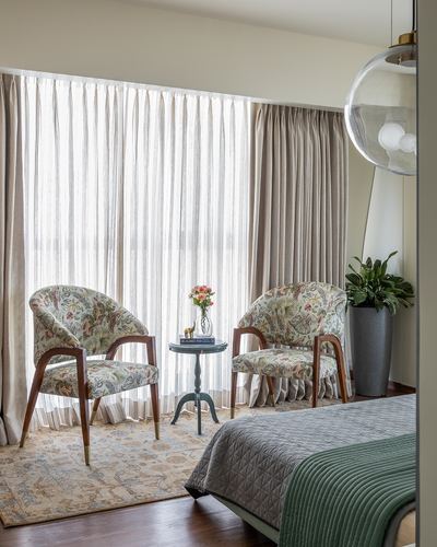 Bedroom corner with floral armchairs, round side table and sheer curtained window in daylight