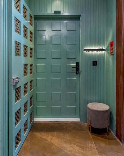 Teal paneled entry door beside jaali screen and round velvet stool on marble floor