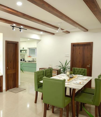 Dining room with green tufted chairs, marble-look table, exposed wood beams, kitchen visible through doorway