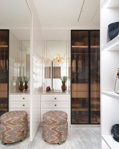 White walk-in closet with mirrored vanity, patterned ottomans, and glass wardrobe doors