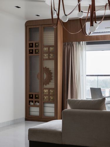Wooden pooja cabinet with carved medallion and brass bells beside a curtained window and beige sofa