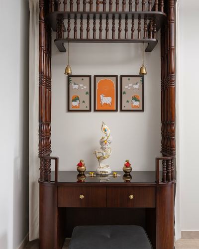 Wooden pooja unit with carved pillars, hanging bells, and framed cow artwork above an idol