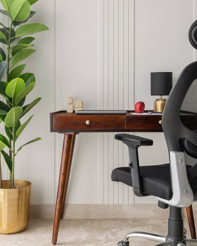 Wooden desk with lamp and figurines beside a black mesh office chair and potted plant