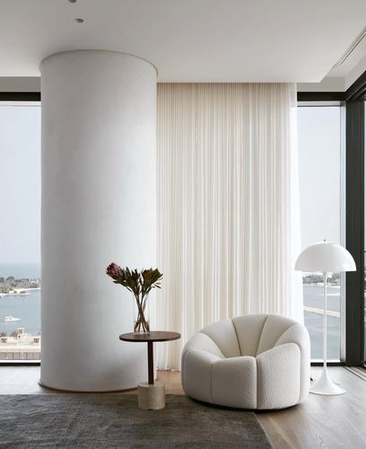 Curved white plaster column beside sheer curtains, boucle armchair, and floor-to-ceiling ocean view windows