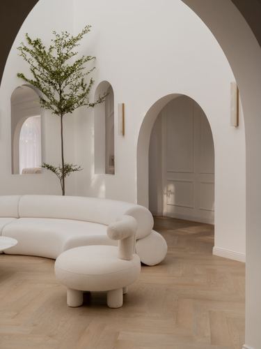Curved boucle sofa and round ottoman in an arched white living room with herringbone wood floor and a small tree