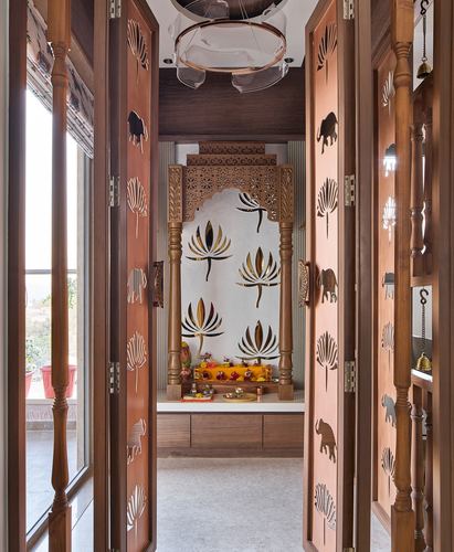View through carved wooden doors into a pooja room with an arched lotus-motif niche and brass details