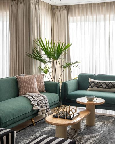 Two green velvet sofas face each other around wood and marble pedestal coffee tables with sheer curtains behind