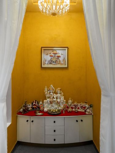 Mustard-walled pooja alcove with white curved cabinet, deity idols, and framed painting under a chandelier