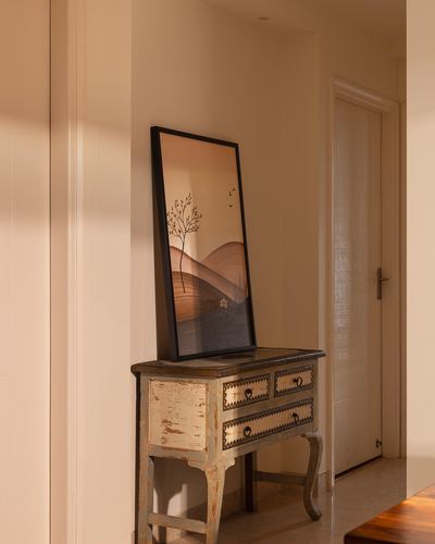 Distressed vintage console table with a leaning framed landscape print in a sunlit beige hallway.