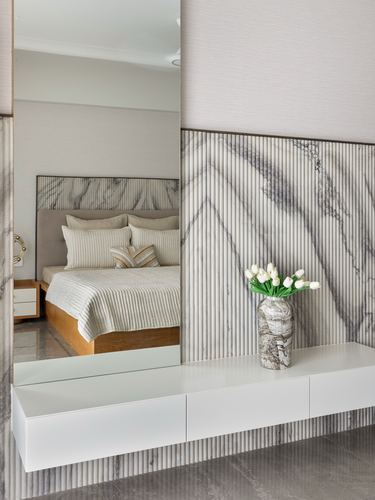 Reflection of a bed in a large mirror beside a fluted marble wall with a floating white console and vase of tulips.