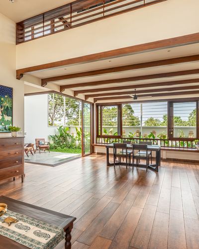Open wood-beamed living and dining space with tall windows opening to greenery in Kozhikode home